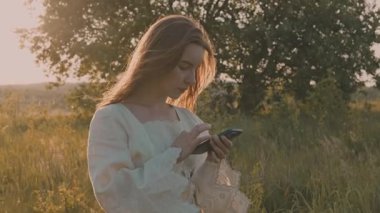 Altın gün batımında kırsal alanda akıllı telefonunu kullanan genç ve çekici bir kadının portresi.