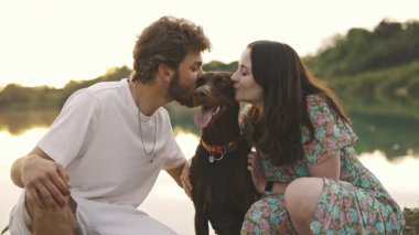 Genç mutlu çift piknik yapıyor ve gün batımında göl kenarında doberman köpekleriyle oynuyorlar.
