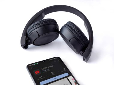 Lod, Israel - July 16,2023: Smartphone with YouTube Kids app play store page and wireless headphones on white background. Top view flat lay with copy space.
