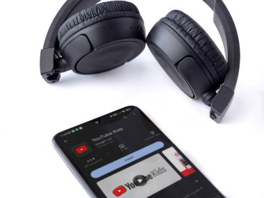 Lod, Israel - July 16,2023: Smartphone with YouTube Kids app play store page and wireless headphones on white background. Top view flat lay with copy space.