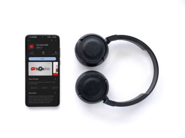 Lod, Israel - July 16,2023: Smartphone with YouTube Kids app play store page and wireless headphones on white background. Top view flat lay with copy space.