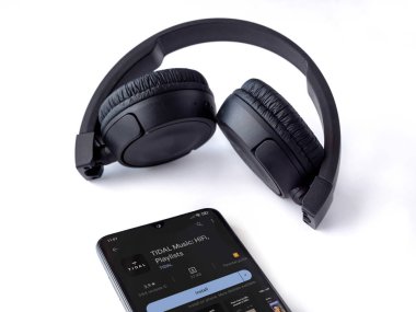 Lod, Israel - July 16,2023: Smartphone with TIDAL Music app play store page and wireless headphones on white background. Top view flat lay with copy space.