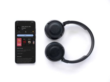 Lod, Israel - July 16,2023: Smartphone with TIDAL Music app play store page and wireless headphones on white background. Top view flat lay with copy space.