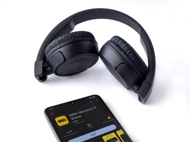 Lod, Israel - July 16,2023: Smartphone with IDMb app play store page and wireless headphones on white background. Top view flat lay with copy space.