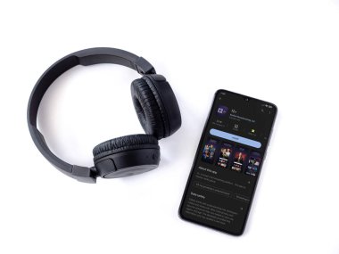 Lod, Israel - July 16,2023: Smartphone with 12+ app play store page and wireless headphones on white background. Top view flat lay with copy space.