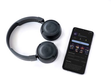 Lod, Israel - July 16,2023: Smartphone with 12+ app play store page and wireless headphones on white background. Top view flat lay with copy space.