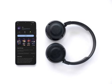 Lod, Israel - July 16,2023: Smartphone with 12+ app play store page and wireless headphones on white background. Top view flat lay with copy space.
