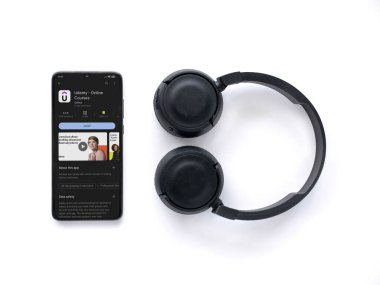 Lod, Israel - July 16,2023: Smartphone with Udemy app play store page and wireless headphones on white background. Top view flat lay with copy space.
