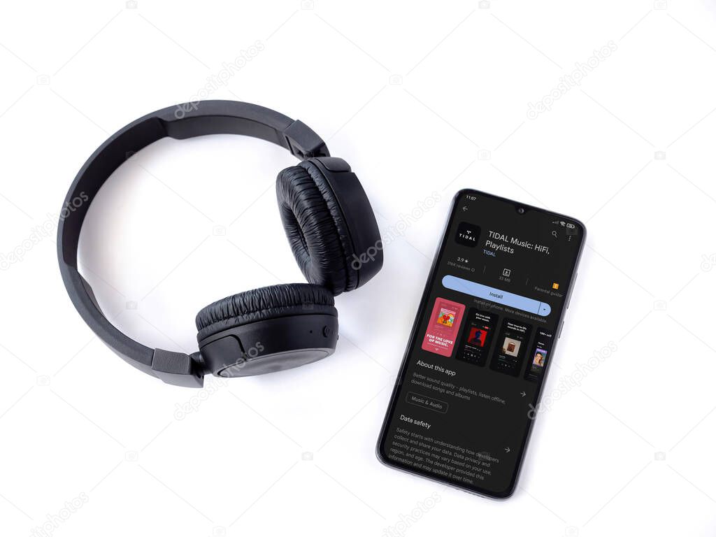 Lod, Israel - July 16,2023: Smartphone with TIDAL Music app play store page and wireless headphones on white background. Top view flat lay with copy space.