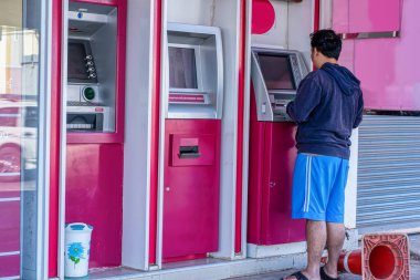 Mavi şortlu ve kapüşonlu bir adam pembe bir bankada ATM kullanıyor..