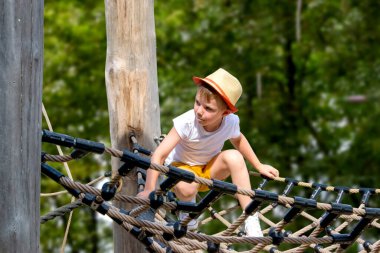 Bir çocuk, sıcak bir yaz gününde parktaki bir çocuk parkına tırmanır. Halk parkındaki çocuk parkı, çocuklar için eğlence ve eğlence, dağcılık eğitimi. Yüksek kalite fotoğraf