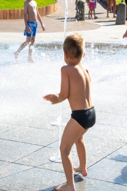 Park çeşmesinde suyla oynayan bir çocuk. Sıcak yaz. Küçük çocuk, güneşli yaz gününde kuru çeşmedeki su jetlerinin arasında oynuyor. Şehirdeki çocuklar için aktif yaz tatili. Yüksek kalite fotoğraf