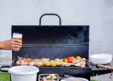 Erkekler ellerinde birayla ızgaranın, sebzelerin ve etin arkasını tutarlar. Seçici odaklanma. Doğa