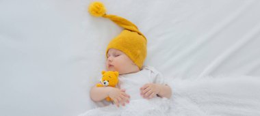 Yeni doğmuş bir bebek beyaz bir arka planda uyuyor, sarı bir ayıya sarılıyor..