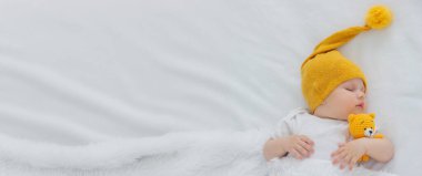 Yeni doğmuş bir bebek beyaz bir arka planda uyuyor, sarı bir ayıya sarılıyor..