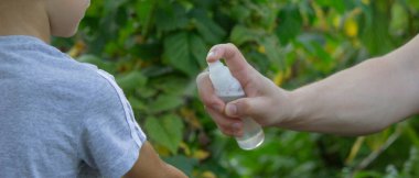 Bir adam parkta, yakın planda oğlunun eline böcek kovucu uygular. Isırık önleyici.