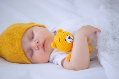 Beyaz elbiseli bir çocuk beyaz bir arka planda uyuyor, küçük örülmüş sarı bir oyuncak ayıya sarılıyor. çocuk