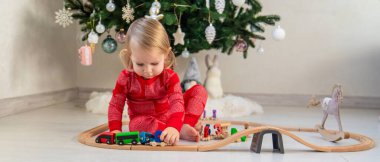 Child in Pajamas Playing with a Wooden Train Set under the Christmas Tree.