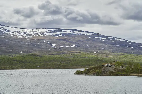 Norveç 'in dağlık kesimlerindeki Jotunheimen gölünün kıyıları boyunca uzanan tipik ahşap ev, kulübe ve arka planda karla kaplı dağlar..