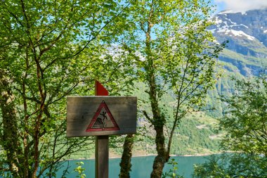 Güneşli bir günde Norveç 'in Geirangerfjord kentindeki derin yeşil ormandan geçen Yedi Kız Kardeş Şelalesi' ne doğru yürüyüş yolu boyunca tehlike işareti..