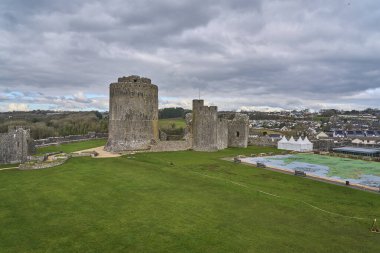 Pembroke, Wales - 02 28 2023: Altında su olan binalarla çevrili yeşil bir tepedeki eski ortaçağ Pembroke kalesi