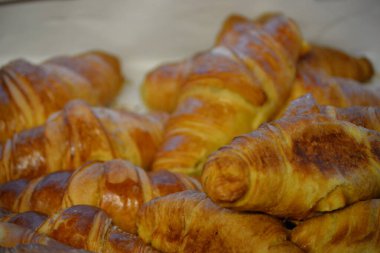 Yeni pişmiş kruvasan yığınının yakın çekimi, altın-kahverengi ve pullu bir yüzeye sahiptirler, ve arka plandaki yumuşak odak ön taraftaki hamur işlerinin çıtır çıtır dokusunu vurgular.