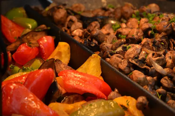 Biri kırmızı ve sarı biber ve domatesler de dahil olmak üzere iki yemek tepsisi, diğeri soteli mantarlar içeriyor, ışıklandırma sıcak, bir pazar veya açık büfe ortamı öneriliyor.