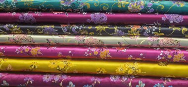 Katlanmış ipek kumaş yığınının canlı bir yakın çekimi, her biri farklı renkte ve süslü çiçek desenli, parlak madde ışığı yansıtıyor ve çeşitli tasarımlar sergiliyor.