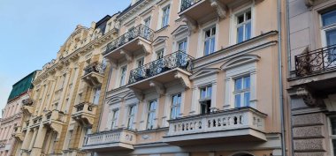 Balkonları ve karmaşık metal parmaklıkları olan eski bir pastel pembe apartman dairesi açık mavi gökyüzünün altında daha süslü, soluk sarı bir binanın yanında..