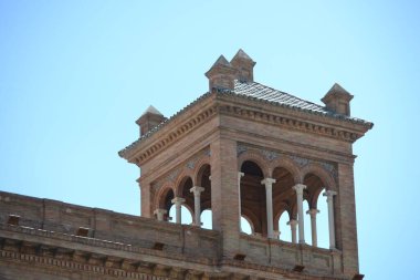Bir fotoğraf, açık mavi gökyüzüne karşı tarihi bir tuğla binanın tepesini gösteriyor. Süslü, küçük kolonlar ve fayanslı bir çatı tarafından desteklenen birden fazla kemer içeren açık hava yapısına sahiptir..