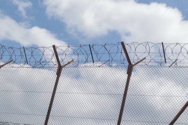 Mavi gökyüzünün arka planında dikenli teller olan metal tel örgüler, beyaz bulutlar. Dikenli teller yakın plan, koruma alanı yüksek metal çitler ve dikenli tellerle çevrili..