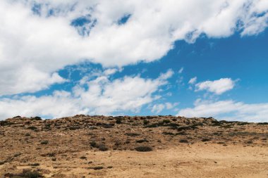 View of sandy, rocky land with white clouds and blue sky in the background. Mountainous nature background for product display with natural sunlight.