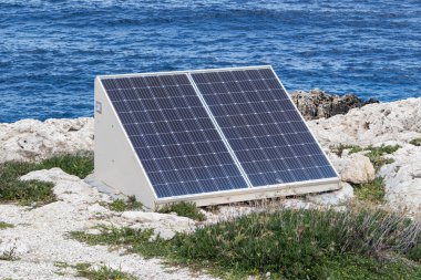 Deniz kıyısındaki kayalıklarda güneş panelleri. Doğa dostu enerji üretimi. Güneşli bir hava, arka planda mavi su olan bir deniz..