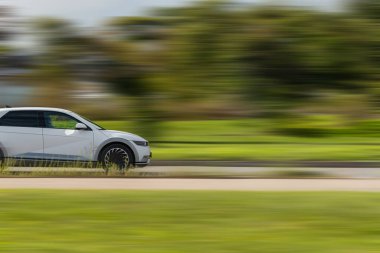 Arka plandaki araç bulanık. Arka planda yeşil çimenler olan beyaz bir araba otoyolda hızlı gidiyor..