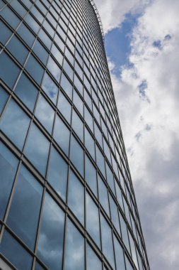 Arka planda mavi, bulutlu gökyüzü olan uzun cam bina duvarının perspektif görüntüsü.