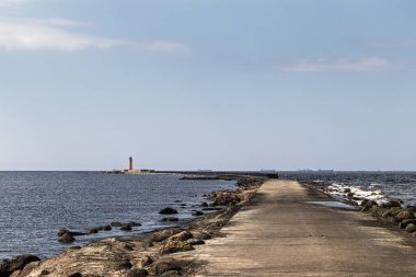Sonunda deniz feneri olan uzun bir beton iskele manzarası. Ufukta büyük kargo gemileri görünüyor..