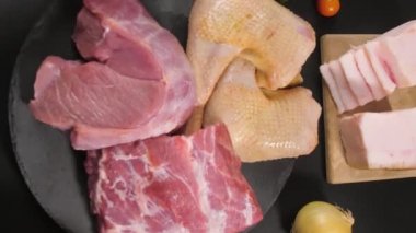 Dikey video. Şefin elleri koyu bir arka plana çiğ dana eti, domuz eti ve tavuk parçaları yerleştirmiş. Etrafta domuz yağı, maydanoz, domates, soğan ve biber var..