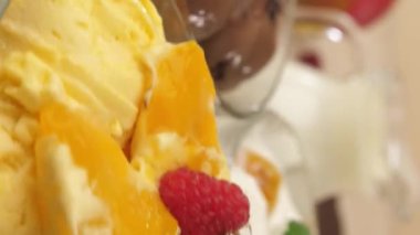 Dikey video. Dondurma, çikolata, mango ve muz aromalı üç cam kase kameranın önünde dönüyor. Sarı dondurmada durur..