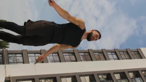 垂直ビデオ 黒い服を着た若い男がスキップロープで屋外にジャンプします 高層ビルを背景に — ストック動画