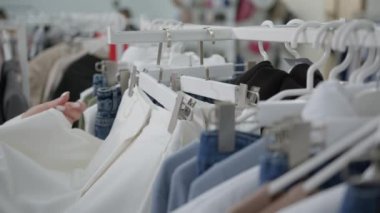 Giysiler mağazadaki askılara asılı. Kadın eli almadan önce beyaz pantolonu seçer. Mağazadan alışveriş..