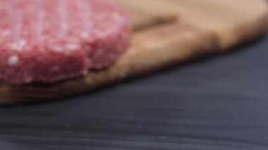 Karanlık bir masa zemininde tahta bir tahtanın üzerinde duran üç çiğ hamburger köftesi boyunca kamera hareketi. Hamburger yapıyorum..