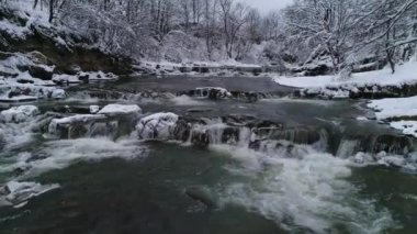 Bulutlu bir günde hızlı akan bir dağ kış nehrinin üzerinde uçmak. Kıyılar karla kaplı..