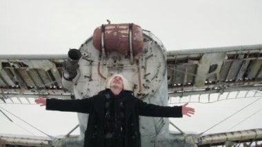 A man in black clothes and a white cap with outstretched arms to the sides sways dreamily against the background of an old Soviet airplane. Snow is flying. Slow motion shooting.