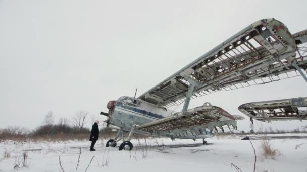 黒い服の男が放棄された壊れたソ連の航空機の前に立っている 翼のように腕を広げる 冬の風景 — ストック動画