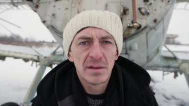 Caucasian man looking forward desolated in sadness. In the background is an old broken plane. Winter scene, snowing. On the head is a white cap and dark clothes.