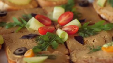 Close-up of rotating sandwiches with pate. On top, slices of cucumber, tomato, olives, dill, arugula.