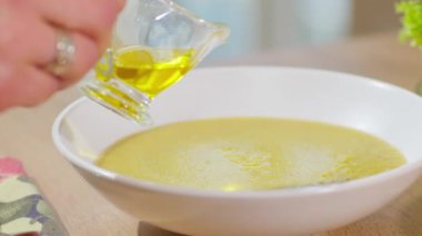 Bir kadın cam bardaktan sıcak krema çorbasına zeytinyağı döker. İlk yemeği pişiriyor ve servis ediyor..