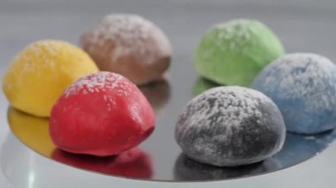 Altı kahverengi, yeşil, kırmızı, siyah, sarı ve siyah mochi kekleri ayna yüzeyinde yatarken dönüyorlar. Hafif bir arkaplanda pasta aromalarının sunumu.