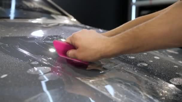 グレーの新しい車のフードにプラスチックスクレーパー接着剤保護フィルムを持つ女性と男性の手 閉めろ 車体保護の概念 — ストック動画