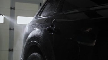 Profesyonel bir işçi, siyah bir SUV 'un sağ arkasını yüksek basınçlı bir yıkama makinesiyle yıkıyor. Aracın parlak yüzeyindeki işçinin yansıması..